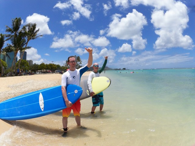 Two guys throwing fist pumps holding their boards, about to start their private surf lesson on a beautiful clear day in Hawaii. Provided by Polu Lani Surf.