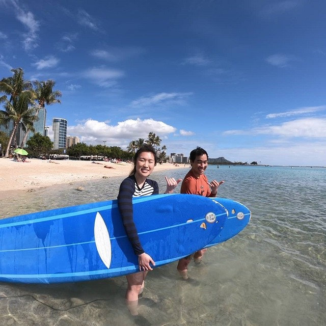A couple with their boards throwing shakas, from the water, about to start their private surf lessons. Provided by Polu Lani Surf.