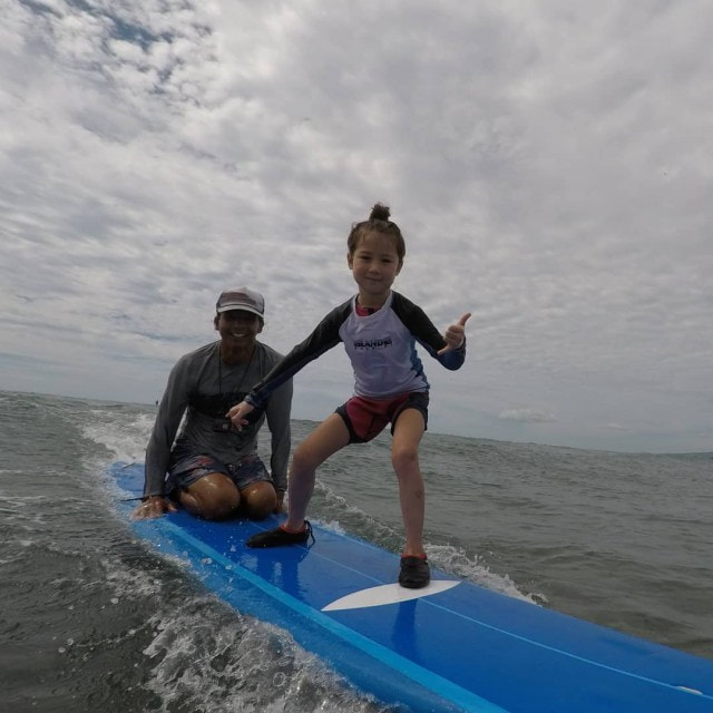 A young girl and instructor catching a wave together, throwing the shaka. Provided by Polu Lani Surf.