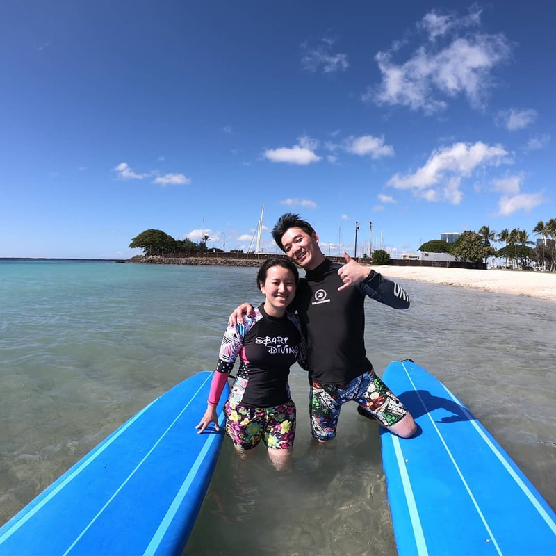 A couple posing together with their boards in the water,  throwing the shaka. Provided by Polu Lani Surf.