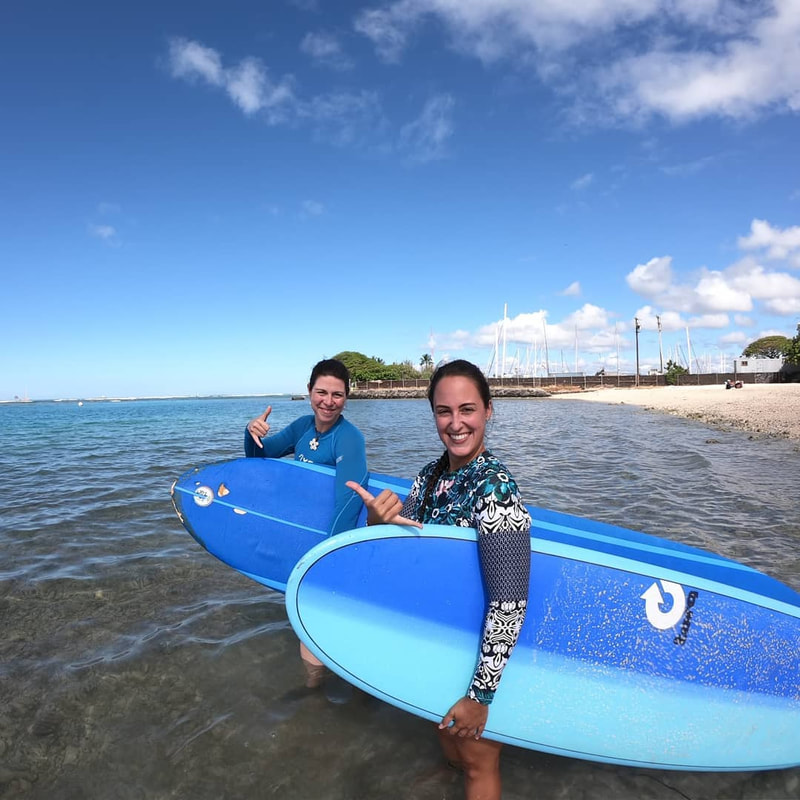 Two young ladies heading out in the water throwing shakas about to start their private surfing lesson. Provided by Polu Lani Surf