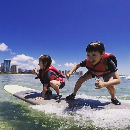 A young girl and boy, riding a wave together while throwing the shaka. Provided by Polu Lani Surf.