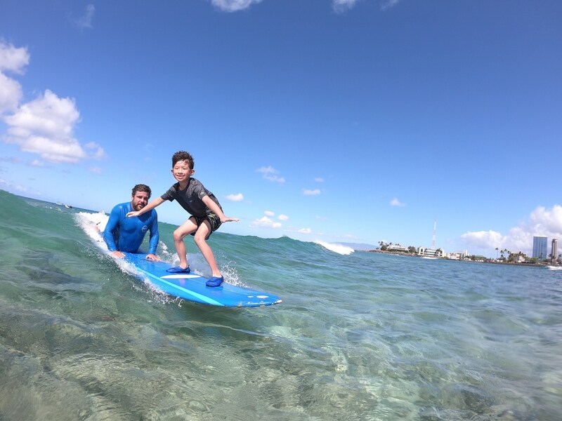 Owner and Instructor, John, and young boy catching a wave together. Able to see the reef through the clear water. Provided by Polu Lani Surf.